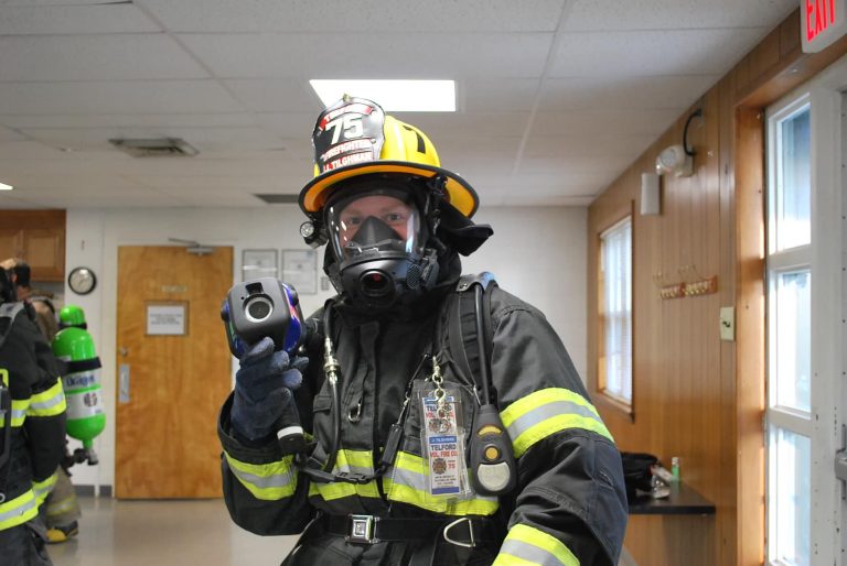 A firefighter dressed in full gear, including a helmet and breathing apparatus, holds a thermal imaging camera. The name tag and ID badge are visible on their chest. The interior setting appears to be a fire station with other firefighting equipment and personnel in the background.