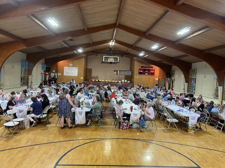 A large gathering takes place in a gymnasium, with rows of tables covered in white tablecloths. People are seated, socializing and eating. The event appears to be well-attended with a festive atmosphere. Banners, signs, and a basketball hoop are visible in the background.