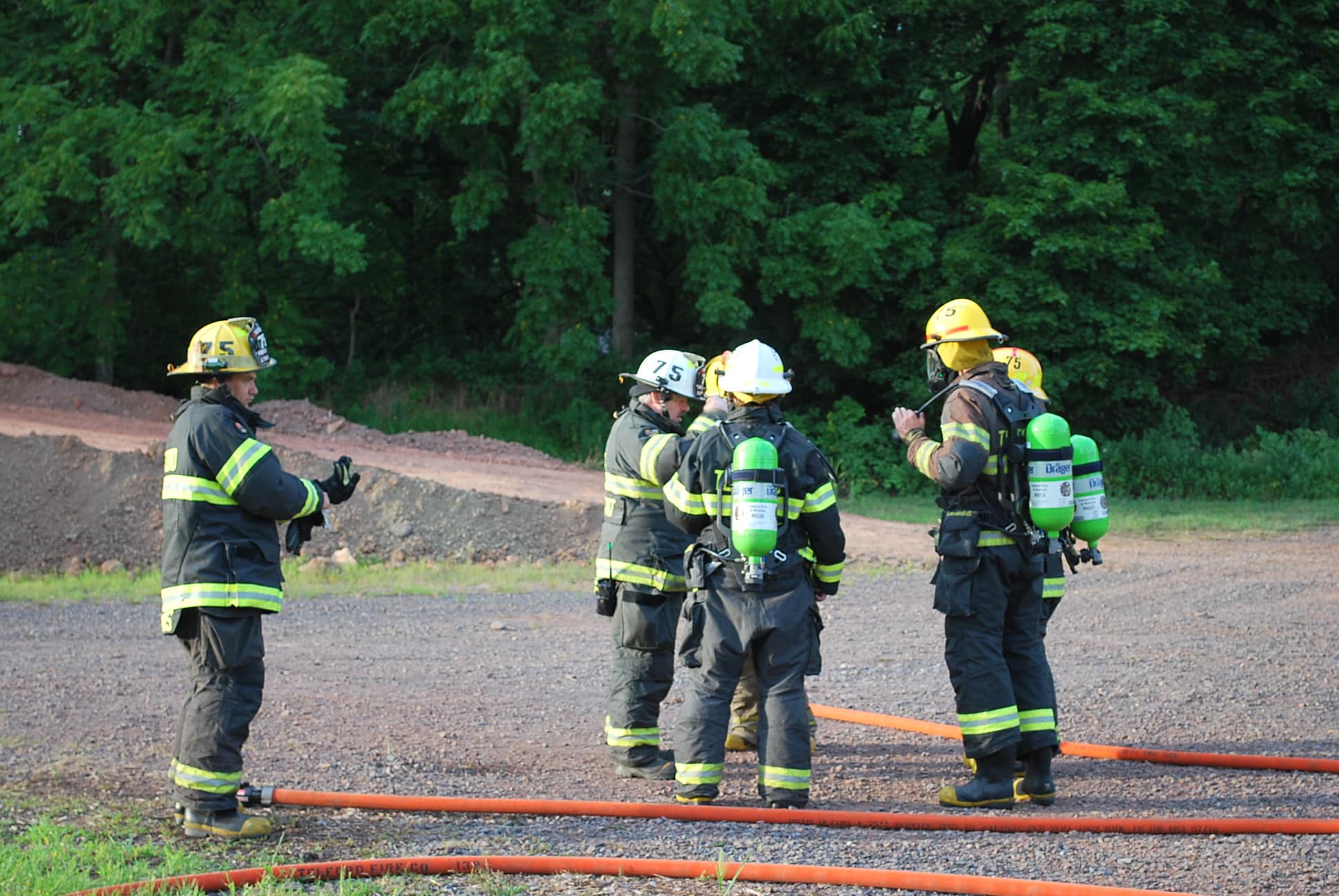 A group of firefighters in full gear, including helmets and oxygen tanks, stand together on a gravel surface with trees in the background. They appear to be in conversation or planning, with fire hoses laid out around them on the ground.