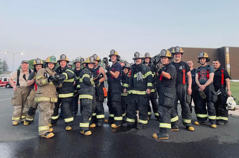 A group of firefighters stands together outside in full gear, some holding equipment. They are posed in front of a building with a fire truck visible in the background. The setting appears to be an urban area, possibly after a drill or a response to an emergency.