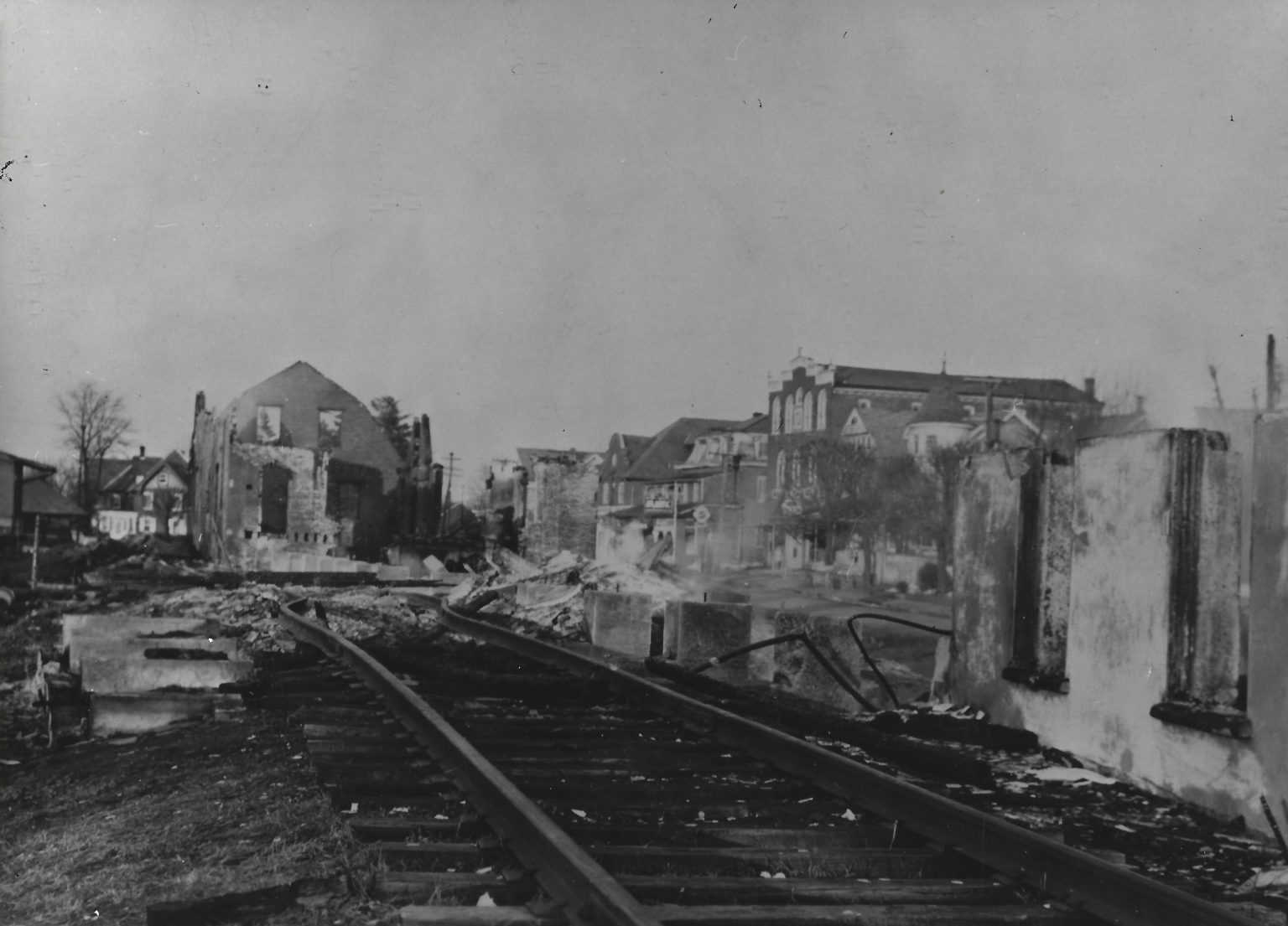 A black-and-white photo shows damaged railroad tracks curving alongside ruined, burnt-out buildings in a town. Smoke rises from debris, and other intact houses are visible in the background.