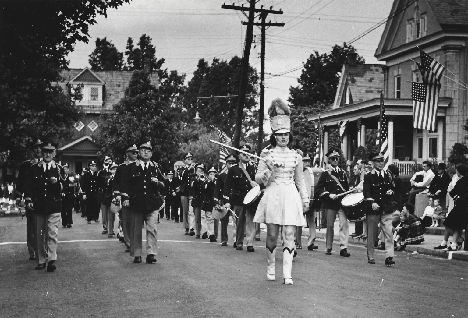 A black-and-white photo of a marching band parading down a residential street, led by a baton twirler in a uniform and feathered hat. Musicians play instruments as bystanders watch from sidewalks and porches, with American flags displayed.