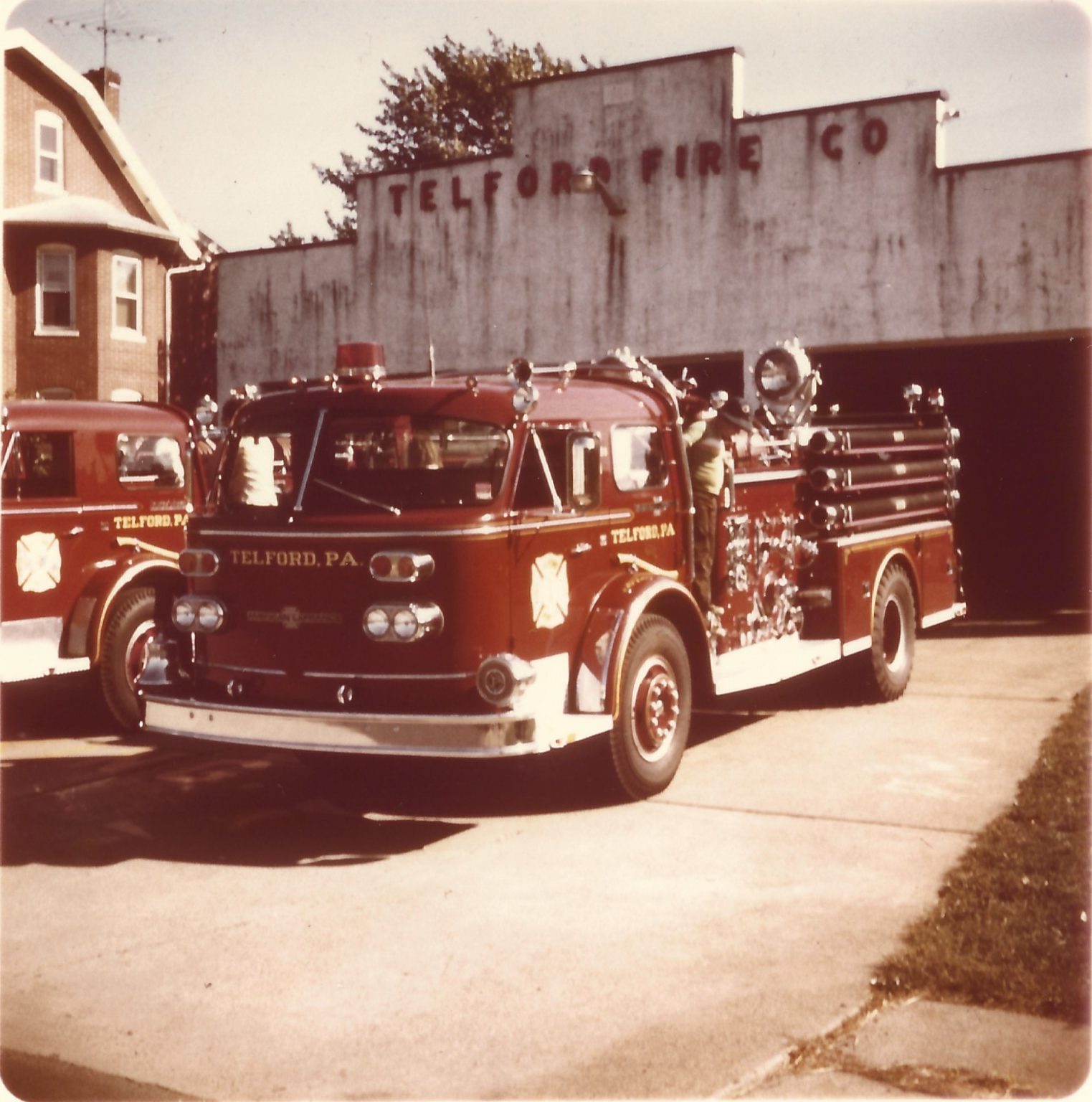 A vintage red fire truck labeled "Telford, PA" is parked outside the Telford Fire Co. station. Another fire truck and a brick house are partially visible in the background. The scene appears to be from decades past.