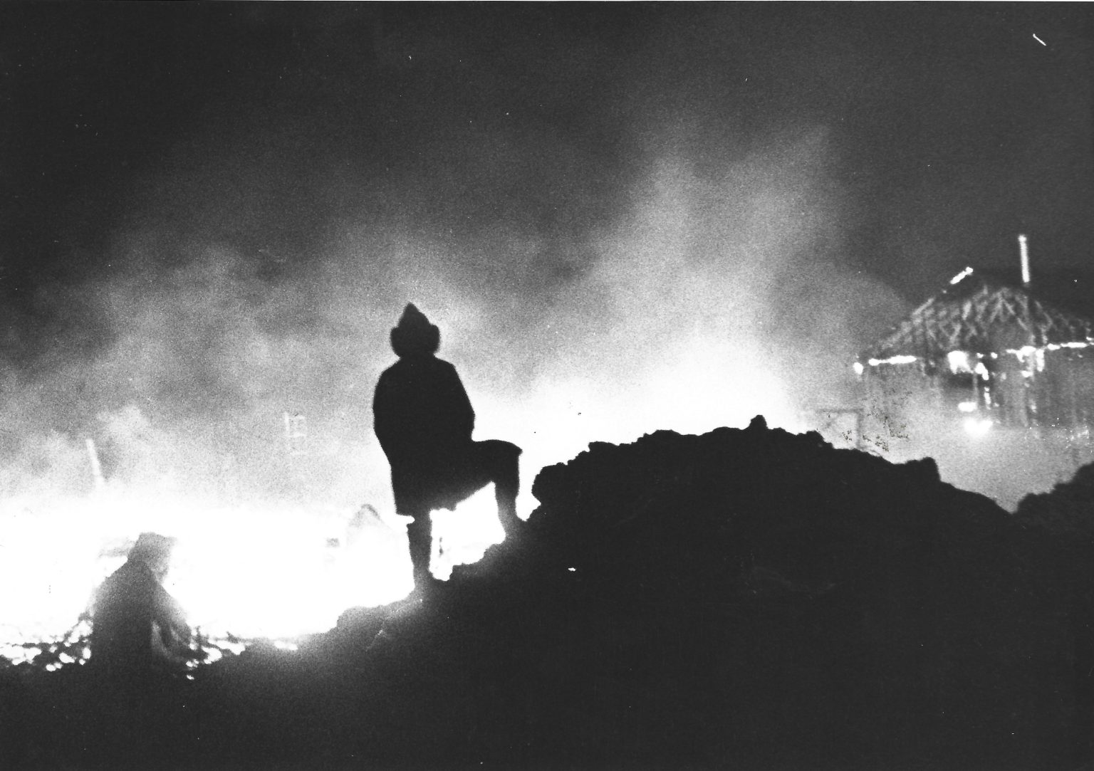 A firefighter in silhouette stands on rubble, looking toward a burning structure at night, with smoke and flames lighting the dark scene. Another figure is visible near the fire.