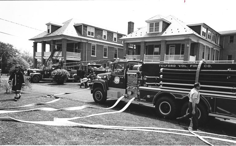 Black and white photo of fire trucks from Telford Volunteer Fire Co. parked between two large brick houses, hoses extended on the ground, with firefighters and civilians nearby.