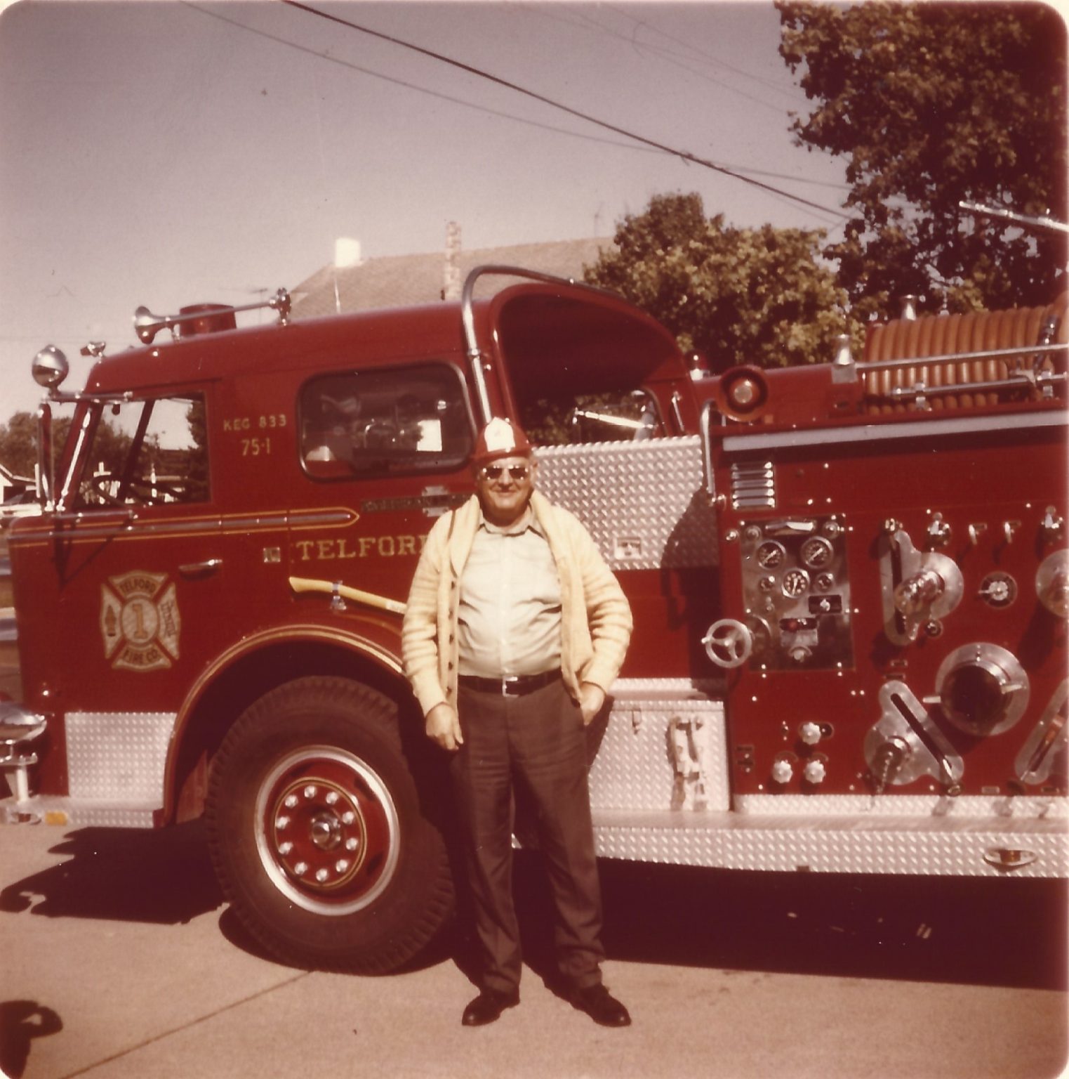 A man wearing a fire helmet stands in front of a red fire truck labeled "Telford." The truck has shiny metal equipment on its side, and trees and power lines are visible in the background.