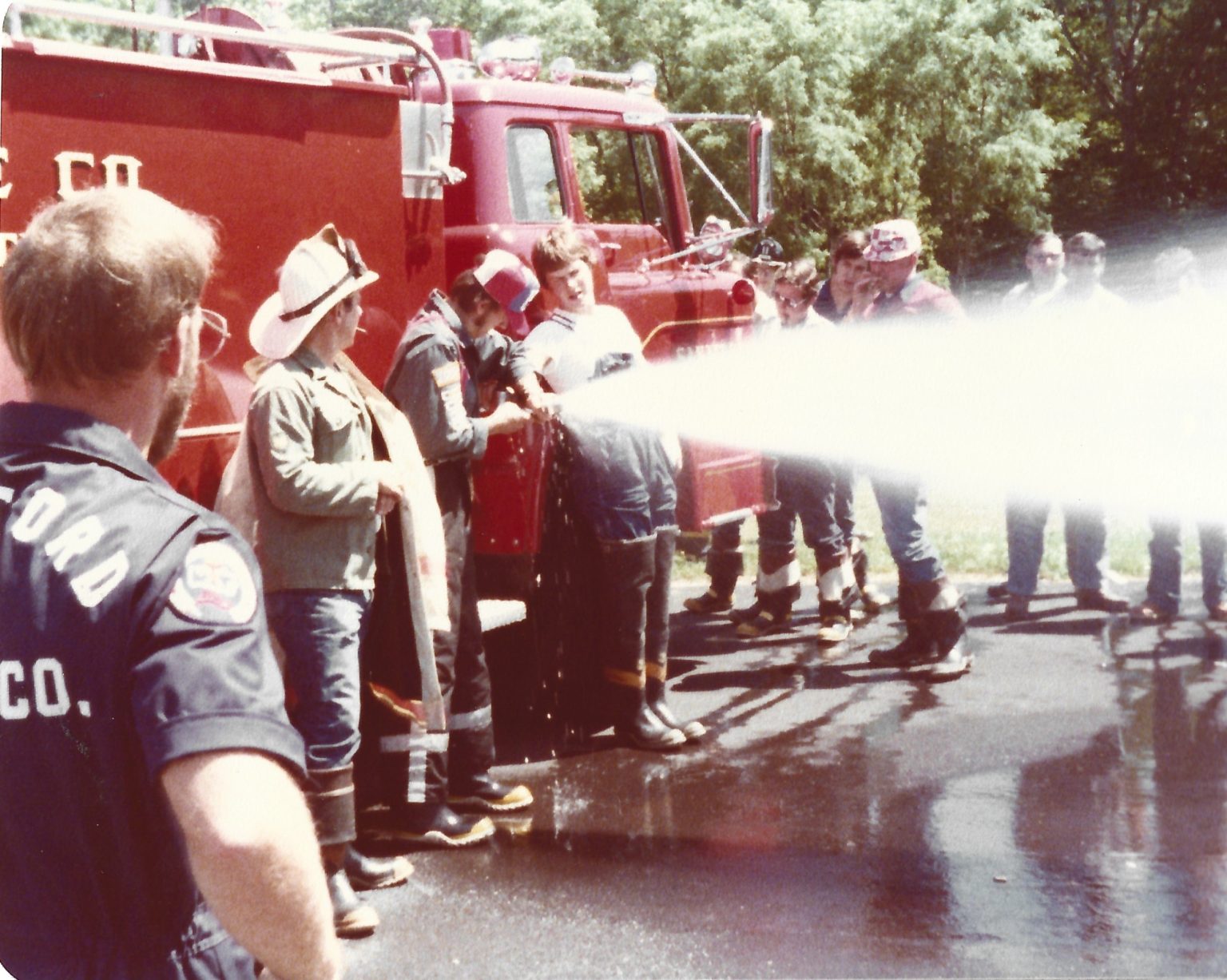 A group of firefighters and trainees, some in helmets and boots, stand by a red fire truck as they spray water from a hose onto wet pavement during a training exercise. Trees are visible in the background.