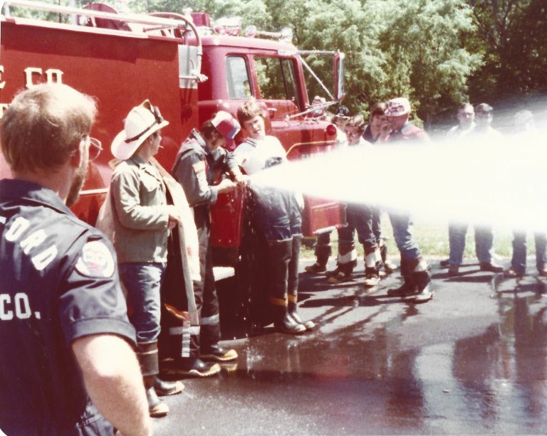 A group of firefighters and trainees, some in helmets and boots, stand by a red fire truck as they spray water from a hose onto wet pavement during a training exercise. Trees are visible in the background.