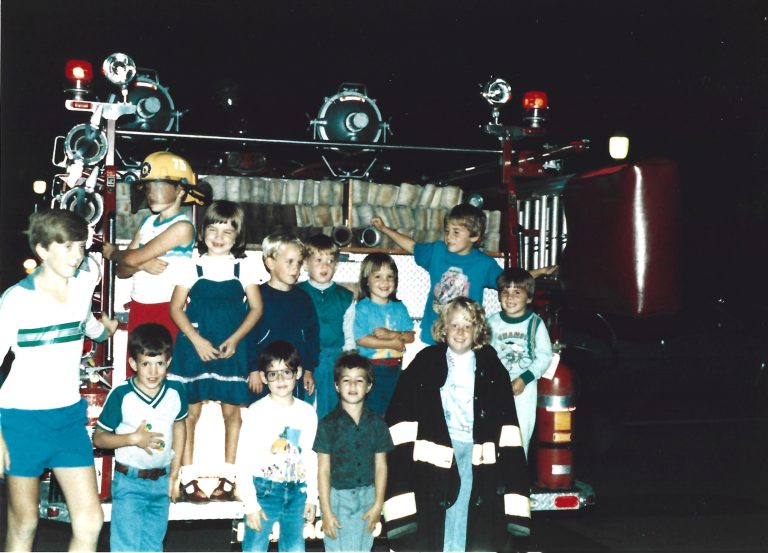 A group of children, some smiling and some posing, stands and sits on the back of a fire truck at night. One child wears a firefighter helmet and another wears an oversized firefighter coat.
