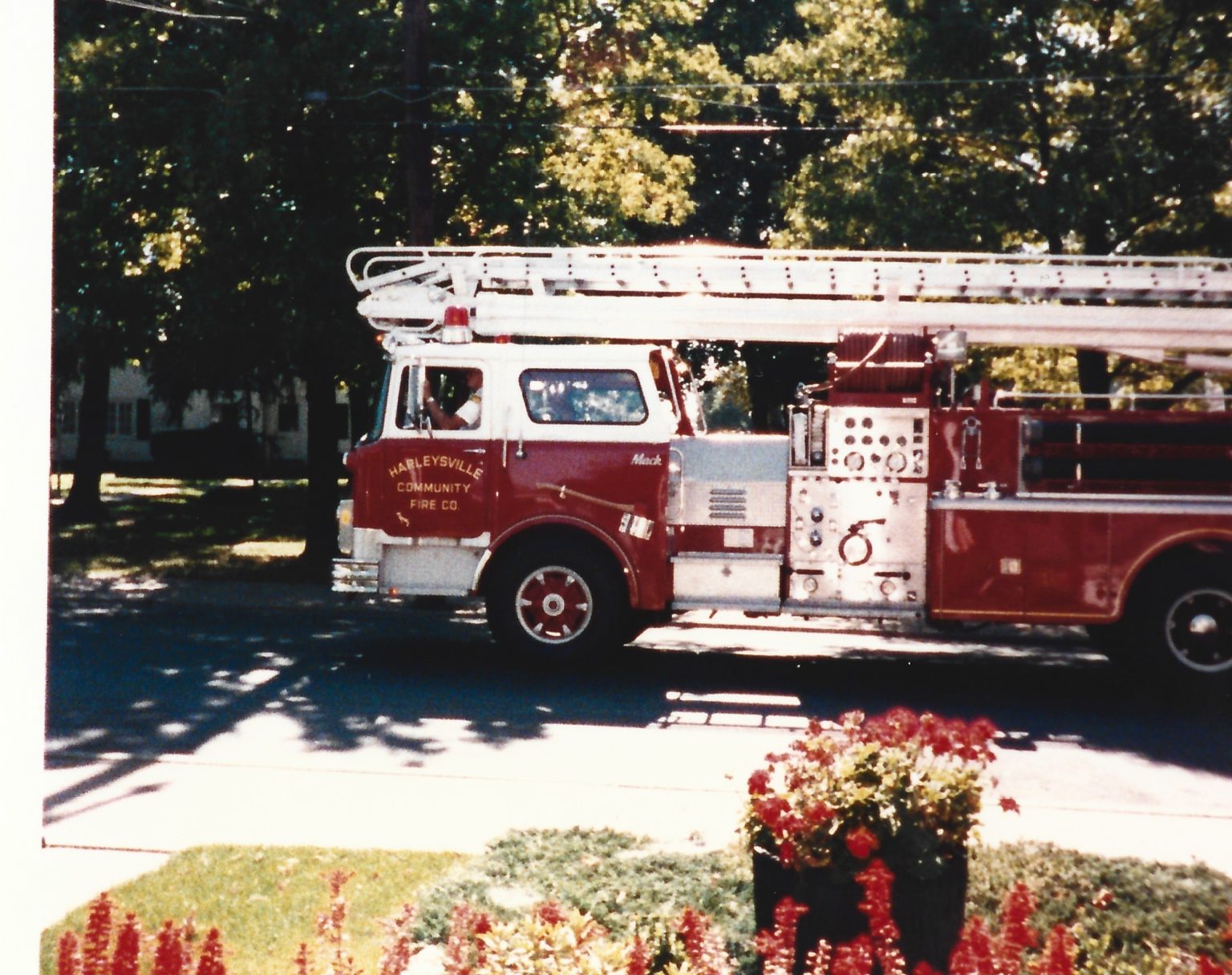 A red fire truck with a long ladder is parked on a shaded street. Trees and houses are in the background, and a flower bed with red and yellow flowers is in the foreground.