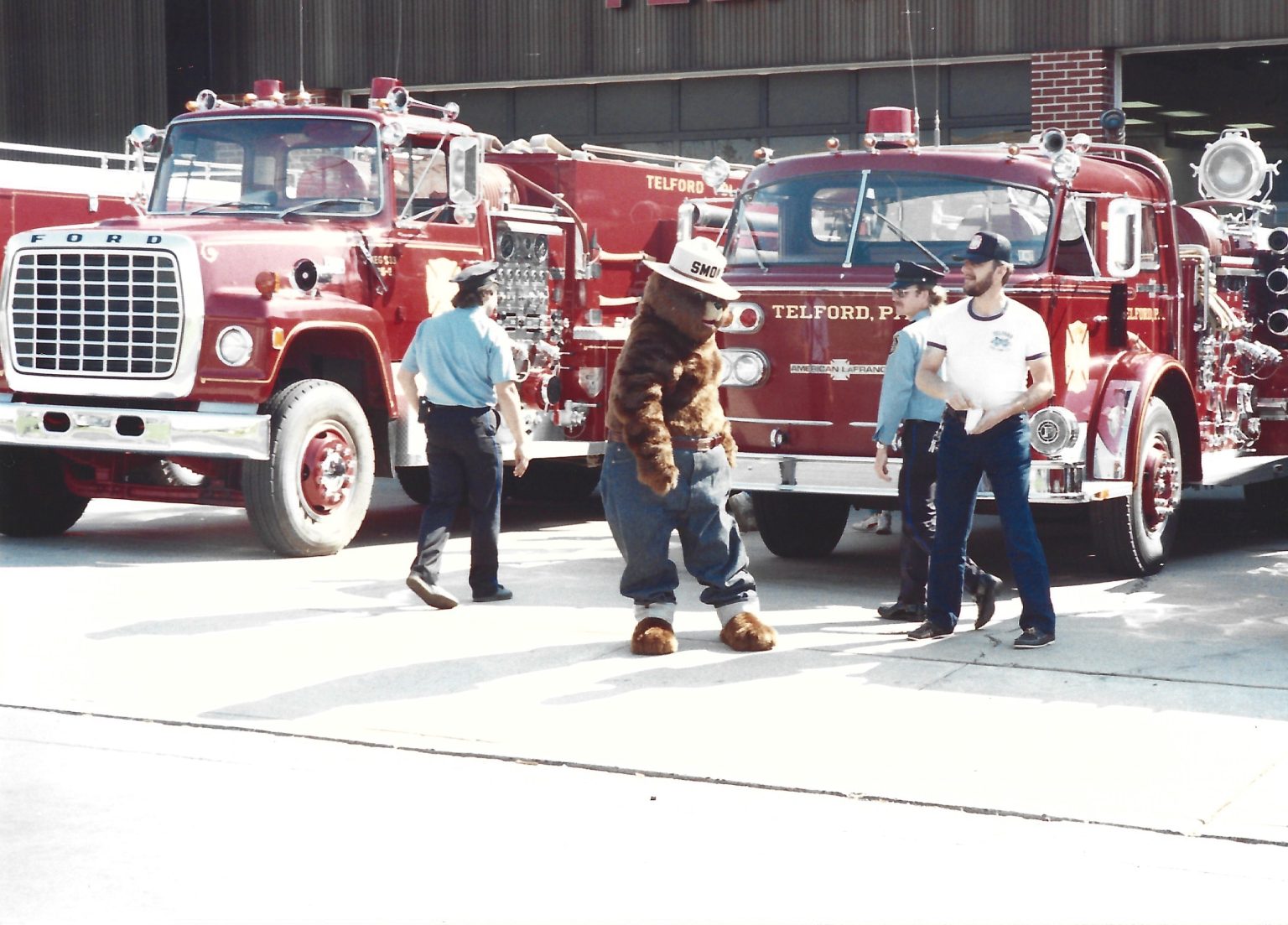 A person in a Smokey Bear costume stands between two vintage red fire trucks with "Telford, PA" on them, alongside three men in casual clothes and uniforms outside a fire station.