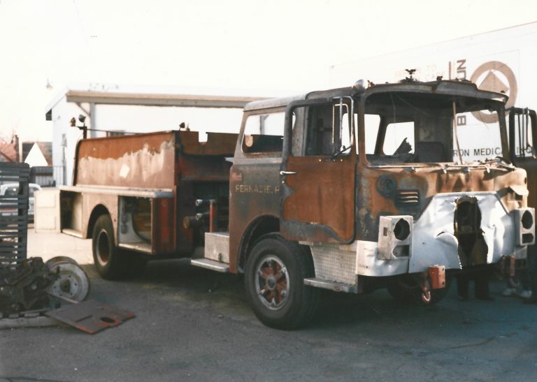 A heavily damaged, rusted fire truck with missing parts is parked in an outdoor lot near a white building and metal debris. The truck’s body is burnt and the cab is partially dismantled.