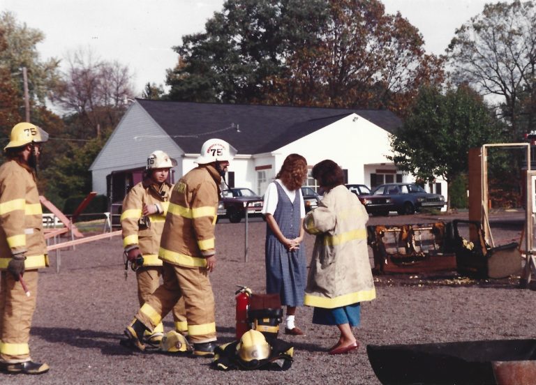 Four firefighters in yellow gear stand near a woman in a blue dress and another woman in a coat, in front of a burned car, with trees and a white house in the background.