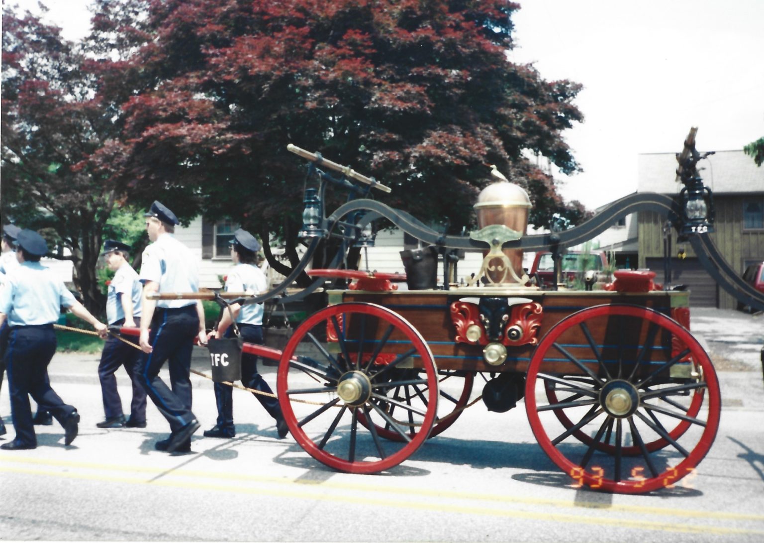 Several people in uniforms pull a vintage horse-drawn fire engine with red wheels and brass fixtures along a street, passing trees and houses on a sunny day.