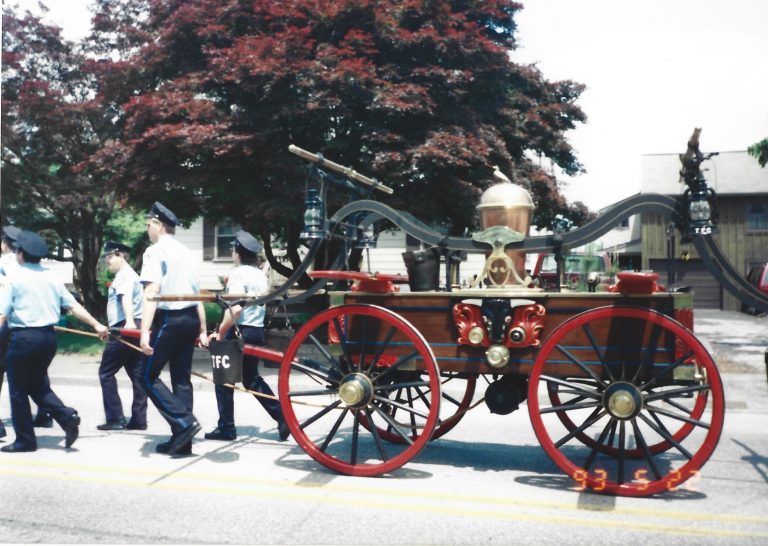 Several people in uniforms pull a vintage horse-drawn fire engine with red wheels and brass fixtures along a street, passing trees and houses on a sunny day.