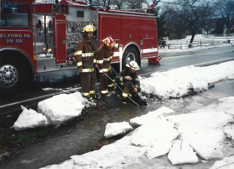 Three firefighters in full gear stand next to a fire truck by a snowy roadside, using tools to clear debris and water from a drain surrounded by melting snow and ice.