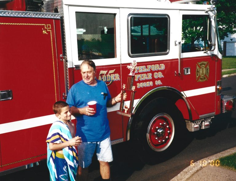 A man holding a red cup stands next to a boy wrapped in a towel in front of a red Telford Fire Co. fire truck on a sunny day. The date "6-24-00" is visible in the bottom right corner.