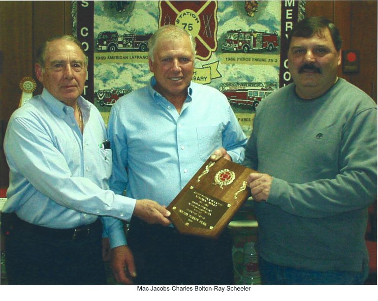 Three men stand indoors; the man in the center holds a wooden plaque with a fire department emblem. A fire truck banner hangs behind them. All are smiling and wearing collared shirts.