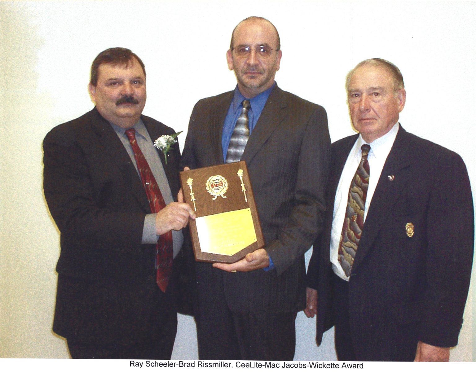 Three men in suits stand together. The man in the middle holds a plaque, while the man on the left gestures toward it. All three are looking at the camera against a plain light-colored wall.