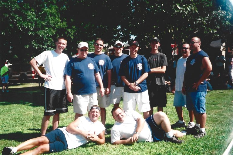 A group of eleven men pose together outside on grass, with nine standing and two lying in front. Some wear matching blue shirts and shorts, while others wear white shirts. It is sunny with trees in the background.