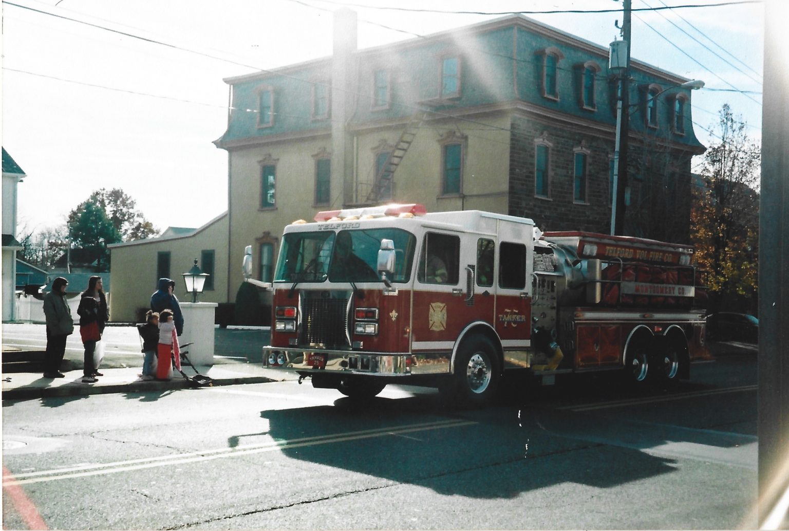 A fire truck is parked on a street in front of a large building. Three adults and a child stand nearby on the sidewalk, next to a fire hydrant, on a sunny day.