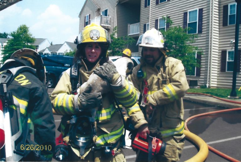 Two firefighters in full gear stand outside an apartment building. One smiles while holding a small dog wrapped in a towel, and the other carries equipment. A fire hose and truck are visible in the background. The date is 6/26/2010.