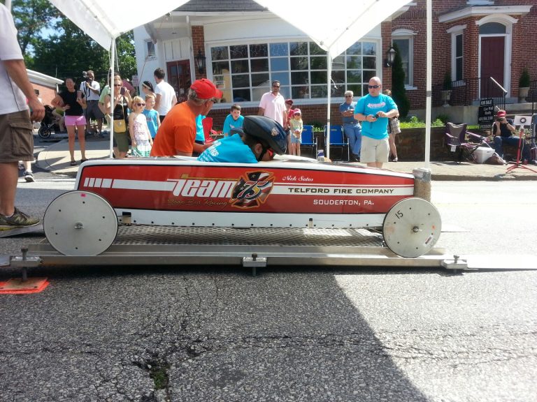 A child wearing a helmet sits in a red and white soapbox derby car labeled "Team 73, Telford Fire Company, Souderton, PA" at the starting line, while adults and other children watch in the background.