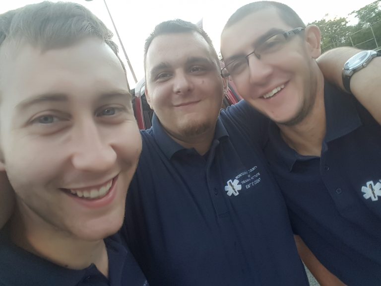 Three young men wearing navy blue shirts with a white medical emblem smile and pose closely together outdoors, appearing happy and friendly.