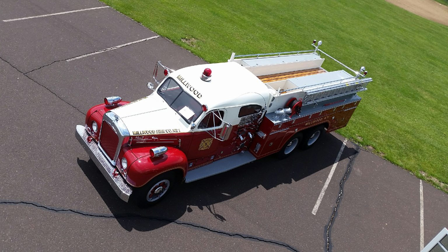 A vintage red and white fire truck is parked on an asphalt lot next to a grassy field, viewed from above. The truck has classic round headlights and silver equipment mounted on its sides.