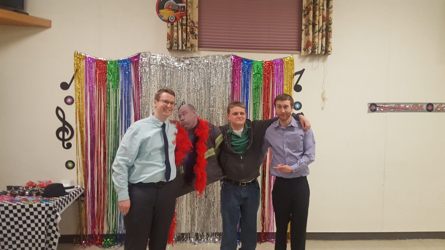 Four men pose in front of a colorful, shiny tinsel backdrop with musical decorations. One man wears a red feather boa and holds a cutout of a person's face. The group appears to be at a party or themed event.