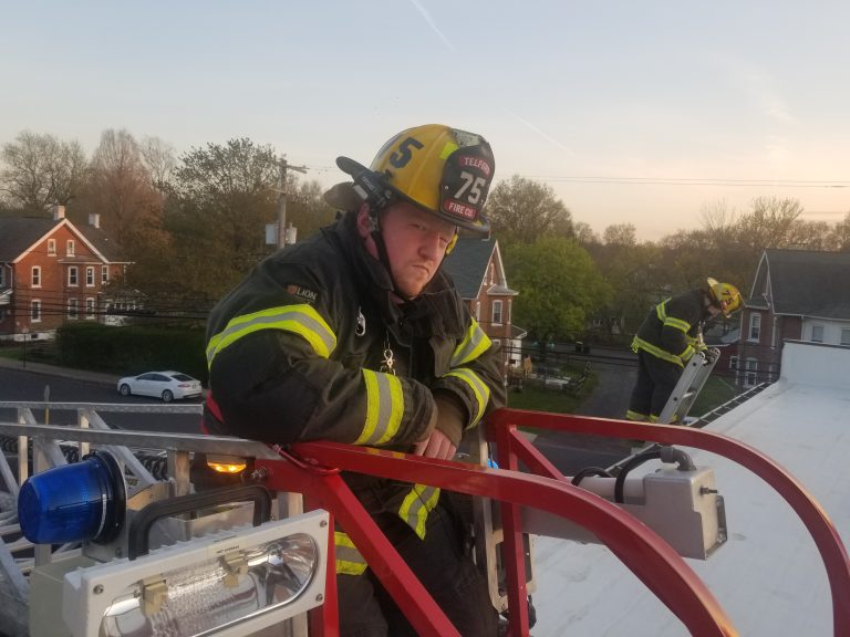 A firefighter wearing a yellow helmet and reflective gear stands on a fire truck’s ladder, looking serious. Another firefighter works nearby on the rooftop. Houses and trees are visible in the background at sunset.