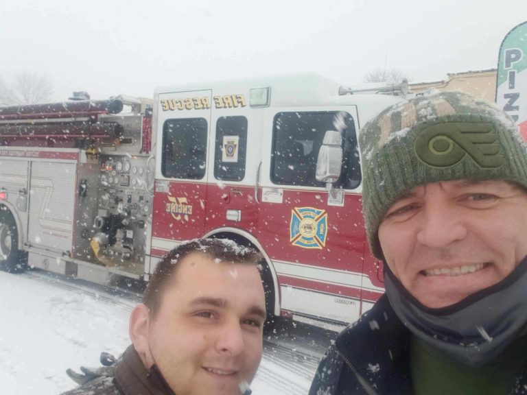 Two men take a selfie outdoors in snowy weather, smiling in front of a red fire rescue truck. Snow covers the ground and the truck, and one man wears a green hat and jacket.