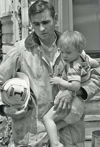 A firefighter in uniform holds a helmet labeled "Chief 1" and carries a young child in his arm. The child clings to him, looking down, as they stand outside a building.