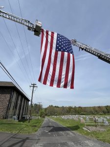 A large American flag hangs vertically between two extended fire truck ladders over a rural road, with a cemetery and trees visible in the background under a clear sky.