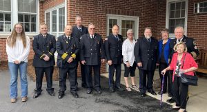 A group of eleven people, including uniformed firefighters and civilians, pose together outside a brick building. Some are smiling and one elderly woman uses a cane. The ground is wet, suggesting recent rain.