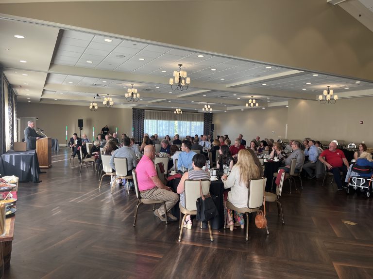 A large group of people sits at round tables in a banquet hall, listening to a speaker at a podium. The room has wooden floors, chandeliers, and large windows letting in daylight.