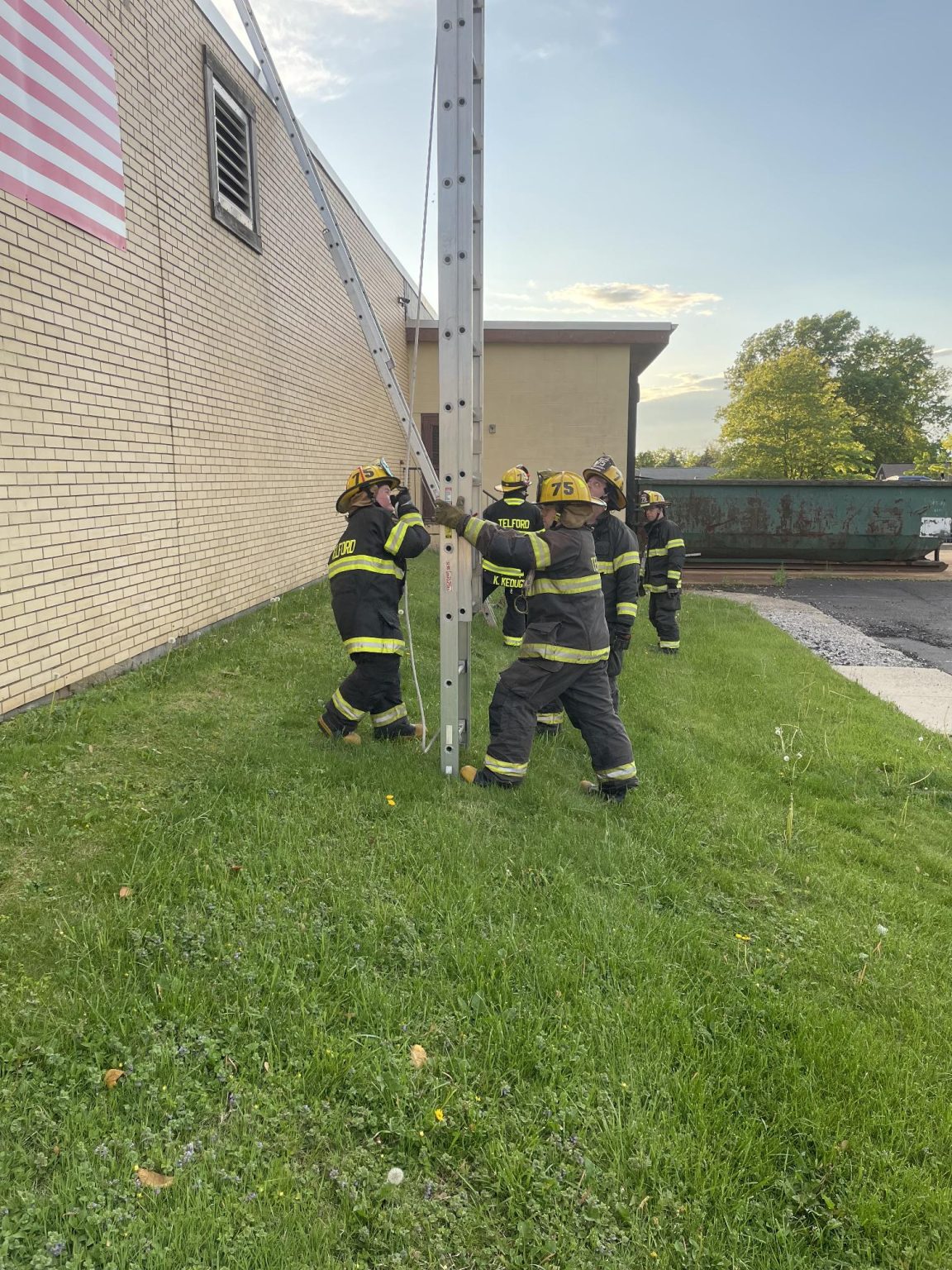 A group of firefighters in full gear work together to stabilize a metal support beam against a brick building on a grassy area, with an American flag and trees in the background.