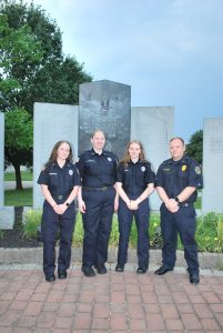 Four uniformed police officers stand in front of a stone memorial monument outdoors, with trees and greenery in the background. They are posing and smiling for the photo.