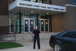 A person in a dark uniform stands in front of the Montgomery County Public Safety Training Campus entrance, with a black car parked nearby and glass doors behind them.