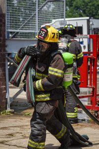 A firefighter in full gear carries a fire hose over their shoulder, while another firefighter works in the background near equipment. Both wear helmets and oxygen tanks. The scene appears to be near a building.