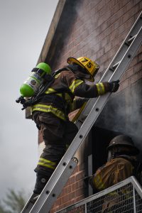 A firefighter in full gear climbs a ladder toward a smoke-filled window of a brick building, while another firefighter stands below, both engaged in an emergency response.