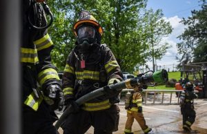 A firefighter in full gear and helmet holds a fire hose, preparing for action. Other firefighters in similar gear are nearby, with trees, equipment, and a blue sky in the background.