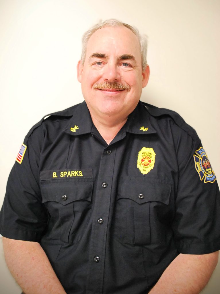 A middle-aged man with short gray hair and a mustache wears a black uniform with fire department badges, a patch on the sleeve, and a name tag reading "B. Sparks." He is smiling and standing against a plain light background.