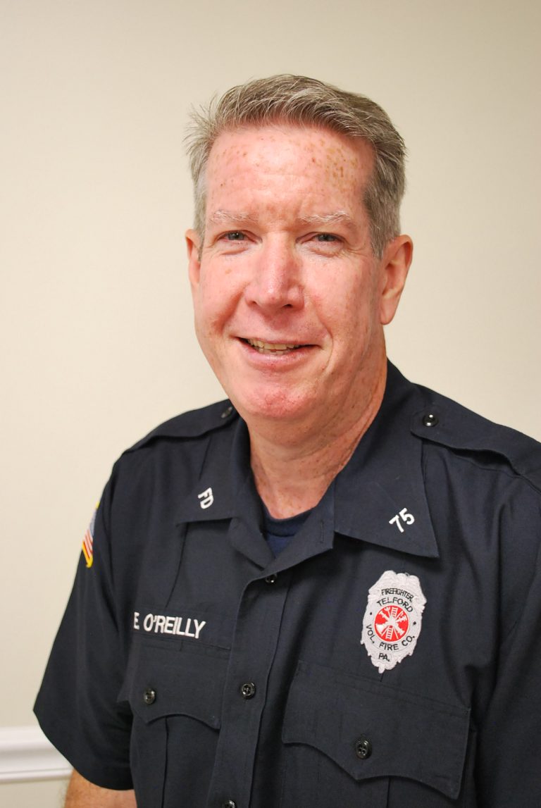A middle-aged man with short gray hair wearing a dark firefighter uniform, smiling softly at the camera. The uniform has a badge reading "Fire Dept" and a patch with an American flag on the sleeve.
