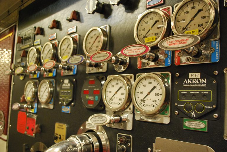 Close-up of a control panel with multiple pressure gauges, dials, switches, and labeled indicators, used for managing and monitoring a fire truck's water system. Labels and color-coded tags are visible.