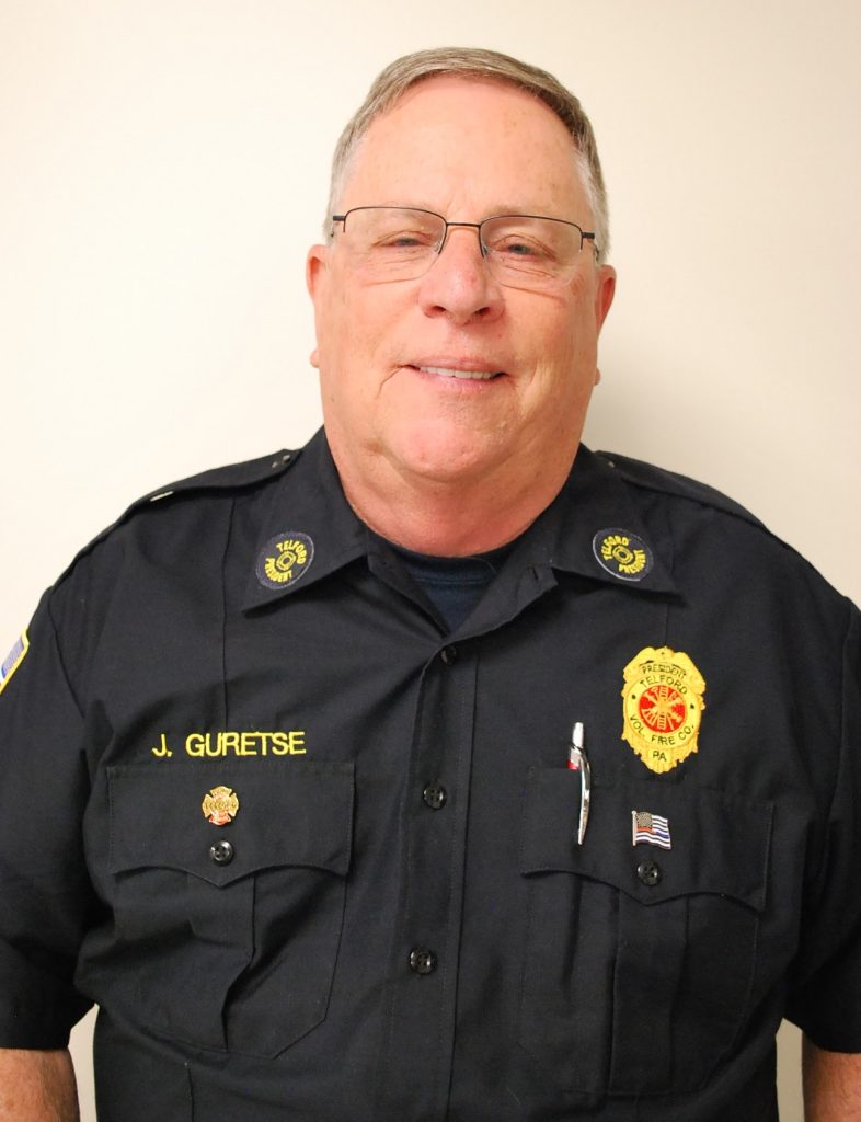 A smiling man wearing glasses and a dark uniform with badges and a name tag that reads "J. GURETSE," standing against a plain light-colored background.