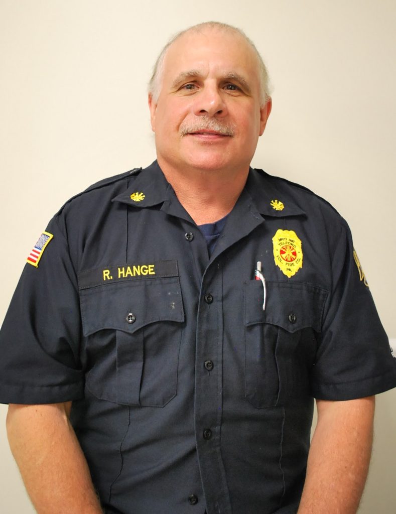A man with short gray hair and a mustache wears a dark blue uniform with badges, an American flag patch, and a name tag reading "R. Hange," standing against a plain light-colored background.
