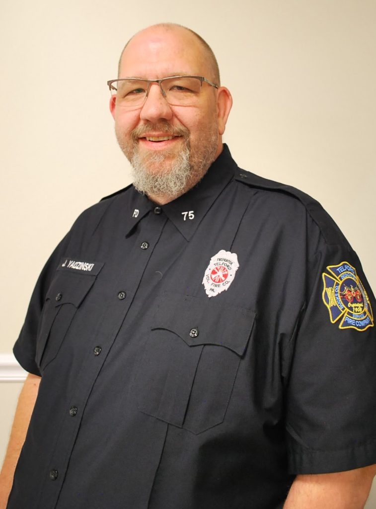 A smiling man with glasses and a beard wears a dark blue fire department uniform with a badge, name tag, and an embroidered patch on the sleeve, standing against a light-colored wall.