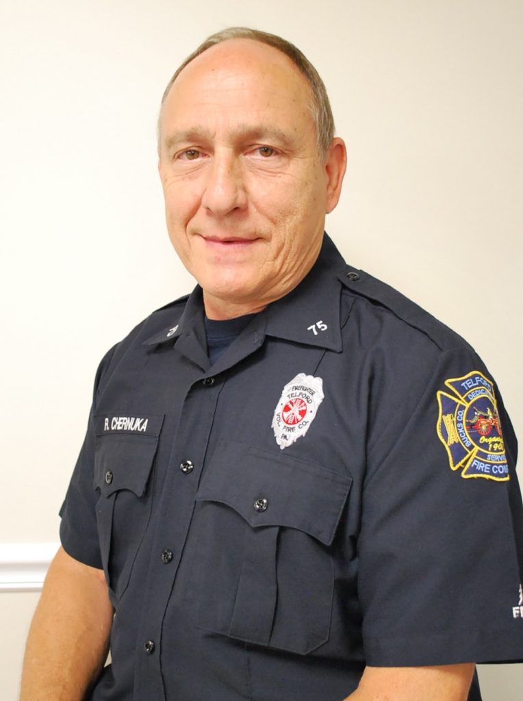 A middle-aged man in a dark fire department uniform with badges and patches is standing against a plain light-colored wall, smiling slightly at the camera.
