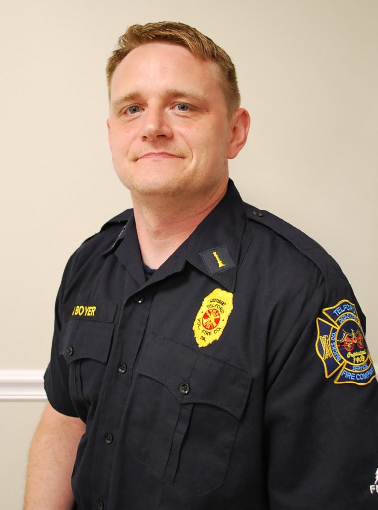 A man in a dark blue fire department uniform with badges and patches is standing against a beige wall, looking slightly to the side and smiling subtly.